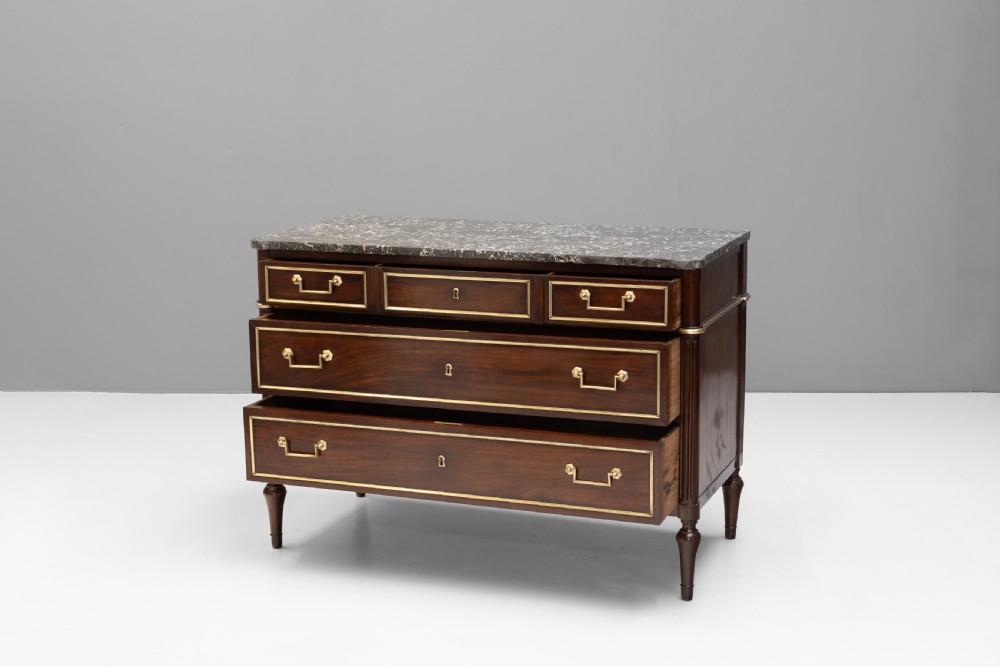 A dark mahogany French Louis XVI style chest of drawers featuring a grey marble top, brass linear moulding, brass swing handles, and turned tapered legs. View III