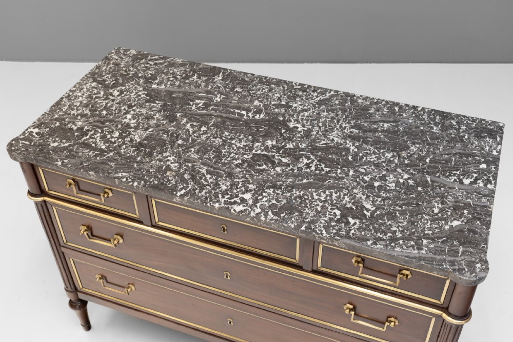 A dark mahogany French Louis XVI style chest of drawers featuring a grey marble top, brass linear moulding, brass swing handles, and turned tapered legs. View IV