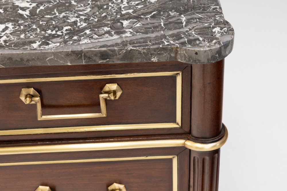 A dark mahogany French Louis XVI style chest of drawers featuring a grey marble top, brass linear moulding, brass swing handles, and turned tapered legs. View VI