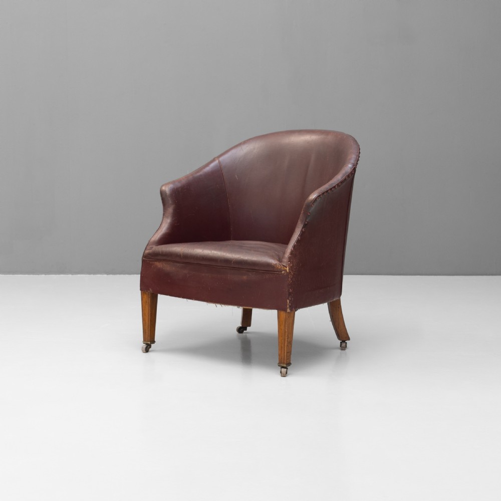 An antique dark brown leather tub chair featuring a rounded backrest, brass studding along the trim, and tapered timber legs on metal castors.