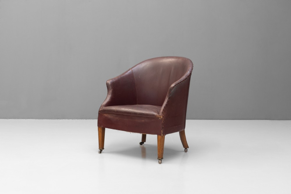 An antique dark brown leather tub chair featuring a rounded backrest, brass studding along the trim, and tapered timber legs on metal castors. View II