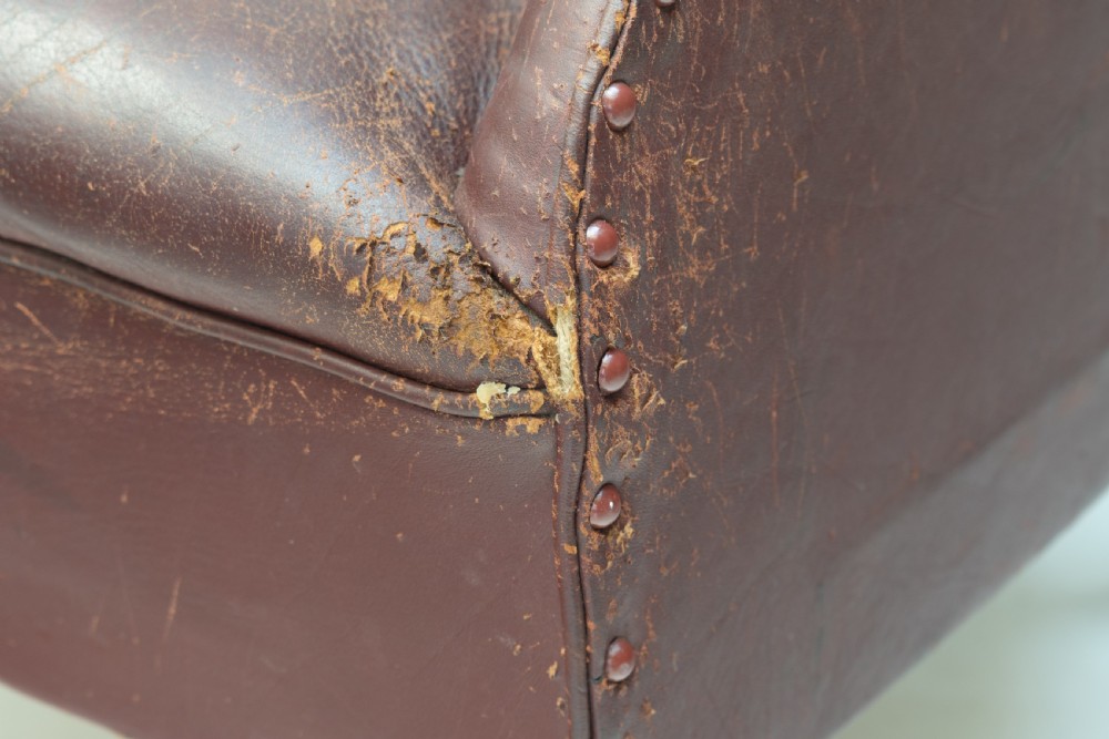 An antique dark brown leather tub chair featuring a rounded backrest, brass studding along the trim, and tapered timber legs on metal castors. View V