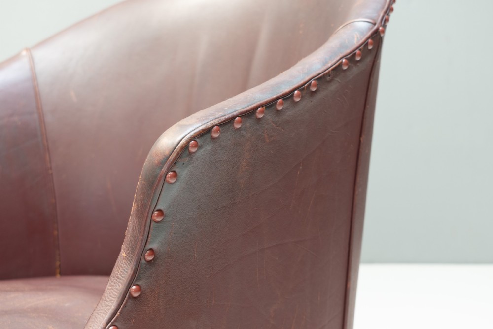 An antique dark brown leather tub chair featuring a rounded backrest, brass studding along the trim, and tapered timber legs on metal castors. View VI