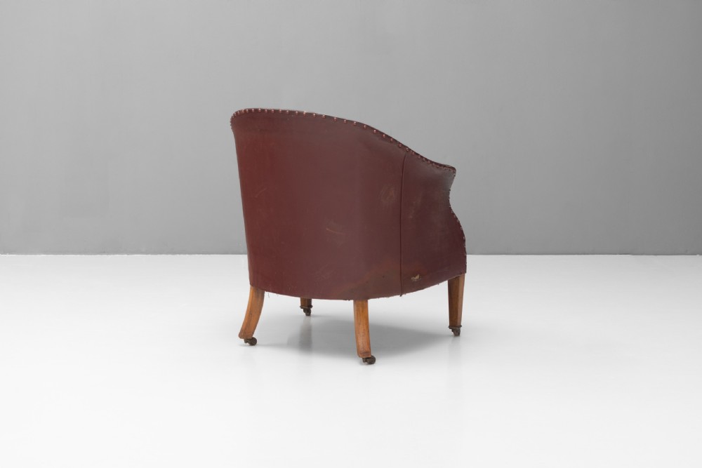 An antique dark brown leather tub chair featuring a rounded backrest, brass studding along the trim, and tapered timber legs on metal castors. View VII
