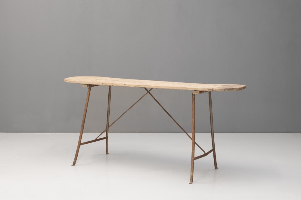 A long narrow rustic table featuring a natural pale wood top with rounded ends supported by slender brown metal legs with diagonal cross-bracing.