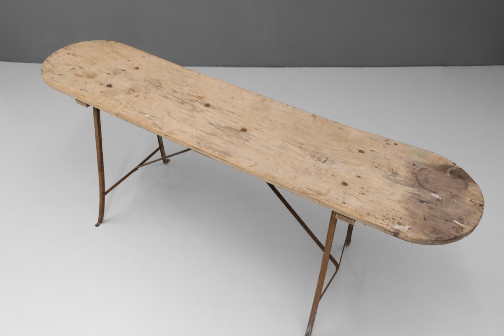 A long narrow rustic table featuring a natural pale wood top with rounded ends supported by slender brown metal legs with diagonal cross-bracing. View VI