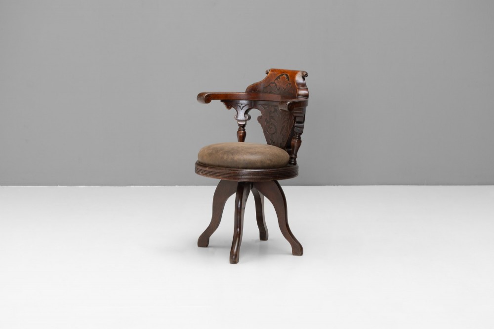 A dark brown Victorian swivel armchair featuring a carved wooden back support and a padded brown leather circular seat.