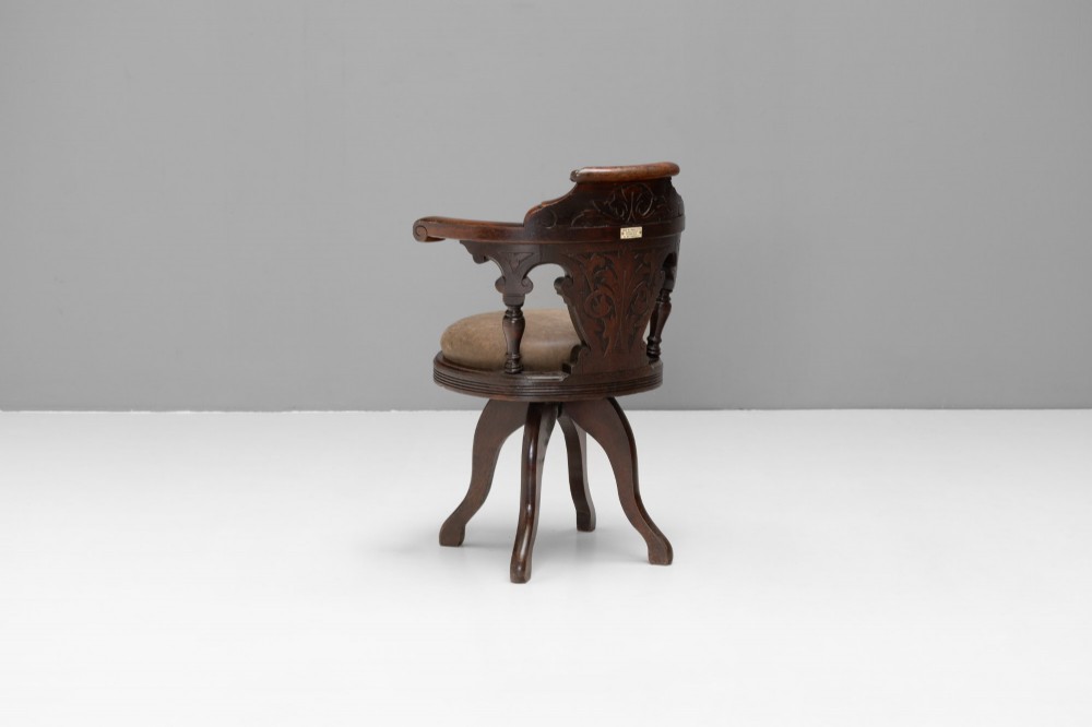 A dark brown Victorian swivel armchair featuring a carved wooden back support and a padded brown leather circular seat. View II