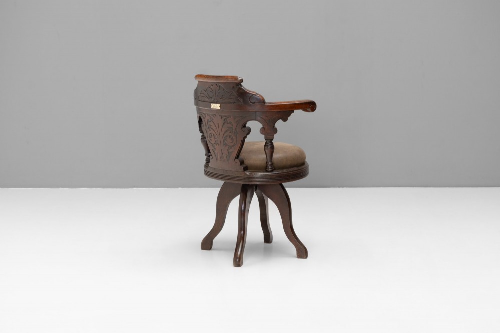 A dark brown Victorian swivel armchair featuring a carved wooden back support and a padded brown leather circular seat. View III