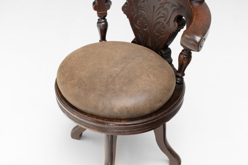 A dark brown Victorian swivel armchair featuring a carved wooden back support and a padded brown leather circular seat. View V