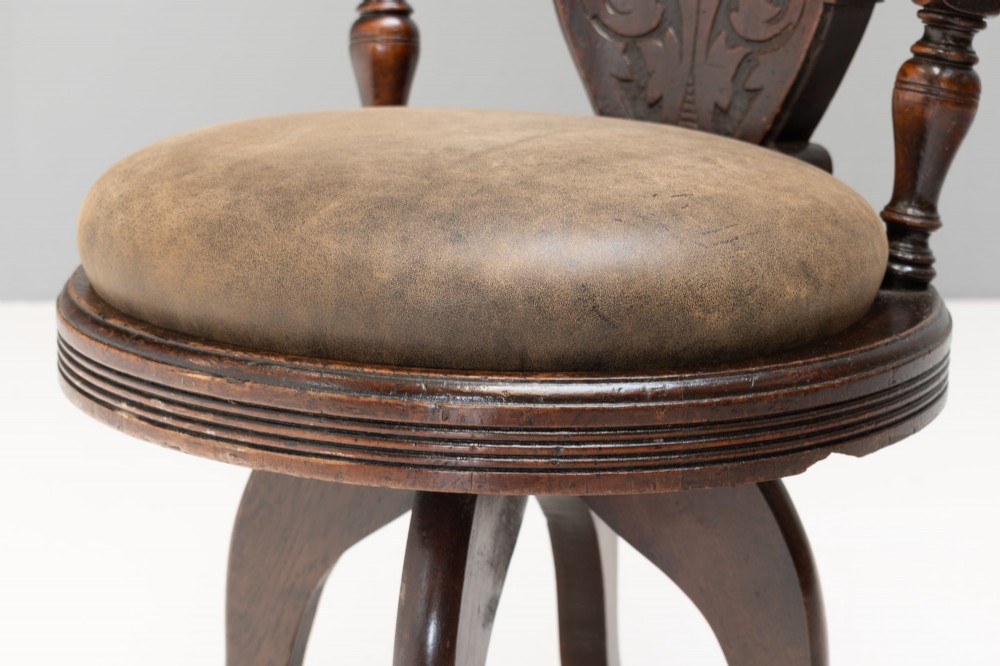 A dark brown Victorian swivel armchair featuring a carved wooden back support and a padded brown leather circular seat. View VII