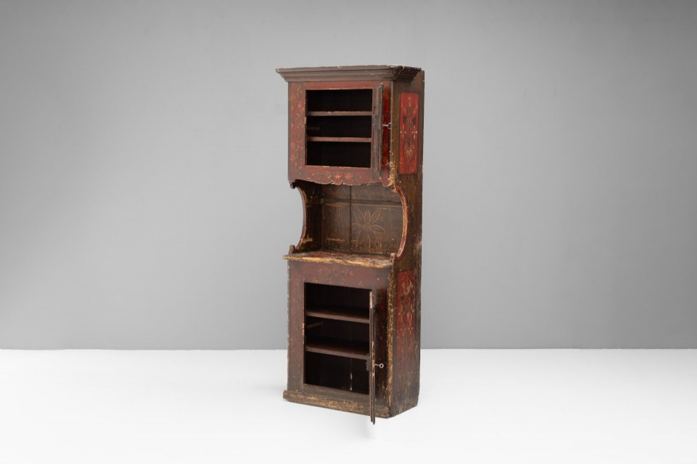 A tall, narrow folk art painted timber cupboard featuring hand-painted floral motifs, date lettering, an upper door, an open central shelf, and a lower door. View II