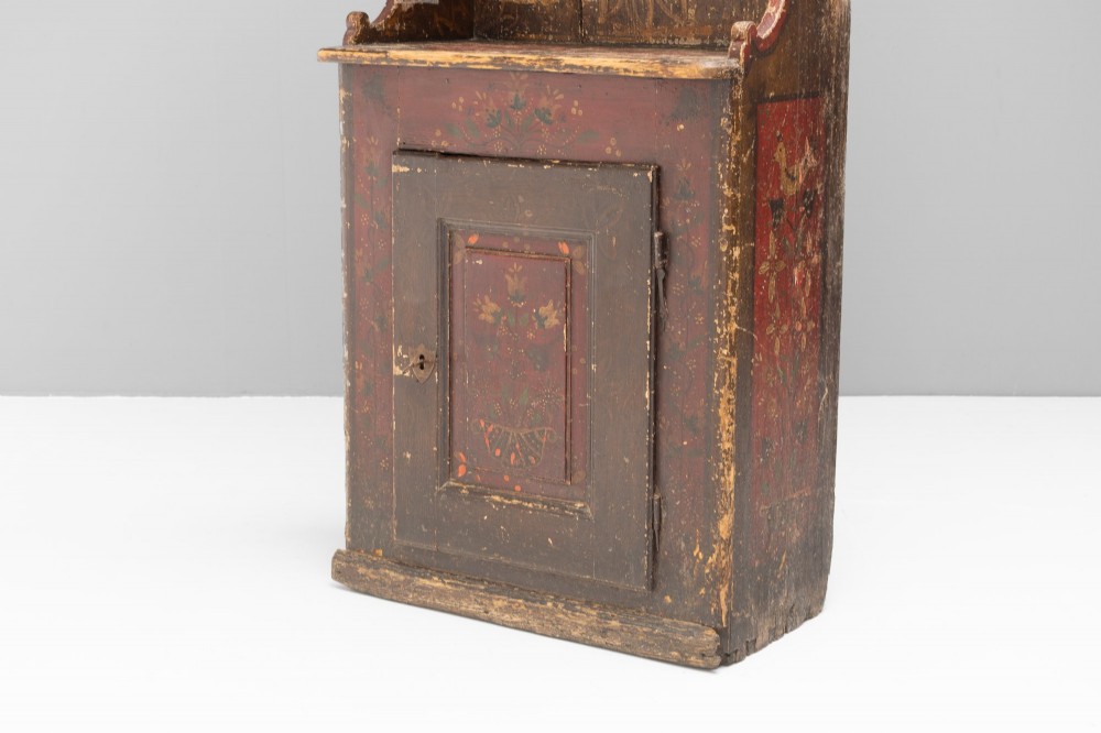 A tall, narrow folk art painted timber cupboard featuring hand-painted floral motifs, date lettering, an upper door, an open central shelf, and a lower door. View VIII