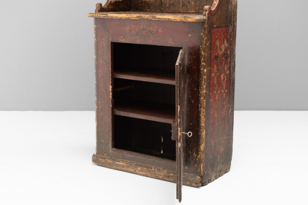 A tall, narrow folk art painted timber cupboard featuring hand-painted floral motifs, date lettering, an upper door, an open central shelf, and a lower door. View IX
