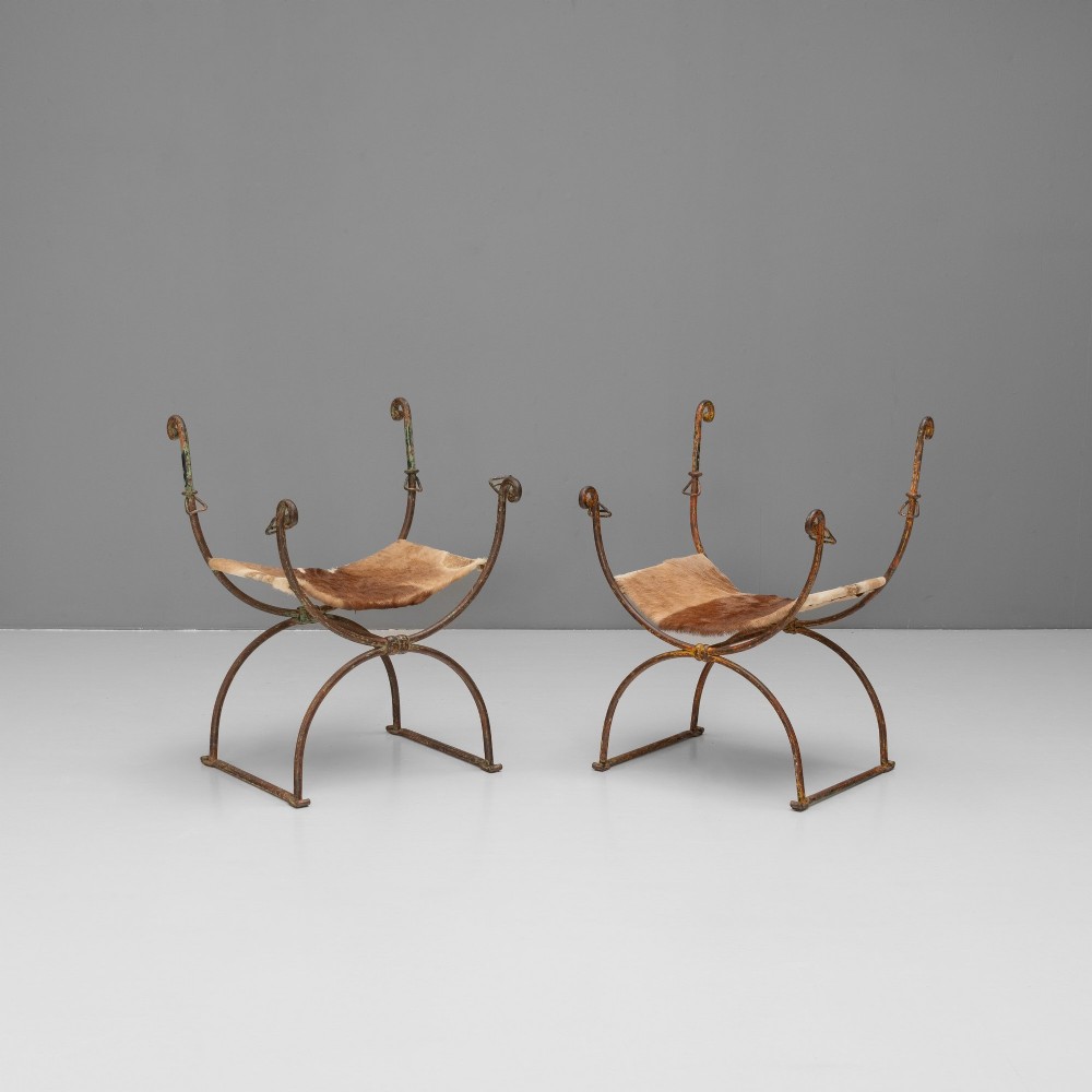 A pair of wrought iron X-frame curule side chairs featuring scrolled metal finials and stretched tan hide sling seats.
