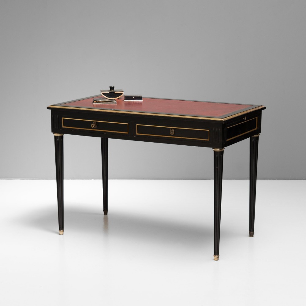 An ebonised wooden writing desk featuring a red leather inset top with gilt brass border inlay, two front drawers, and tapered fluted legs.
