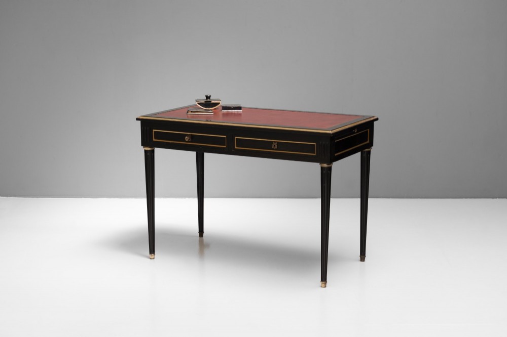 An ebonised wooden writing desk featuring a red leather inset top with gilt brass border inlay, two front drawers, and tapered fluted legs. View II