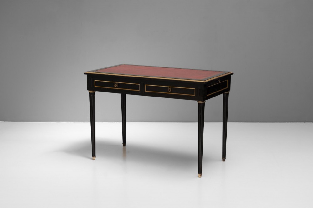 An ebonised wooden writing desk featuring a red leather inset top with gilt brass border inlay, two front drawers, and tapered fluted legs. View III