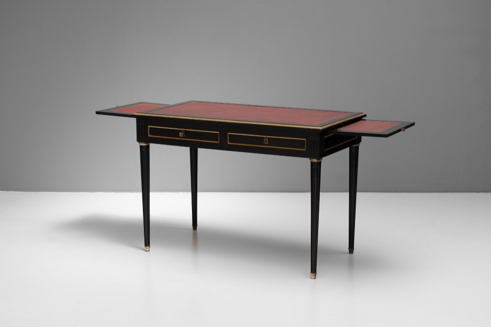 An ebonised wooden writing desk featuring a red leather inset top with gilt brass border inlay, two front drawers, and tapered fluted legs. View IV