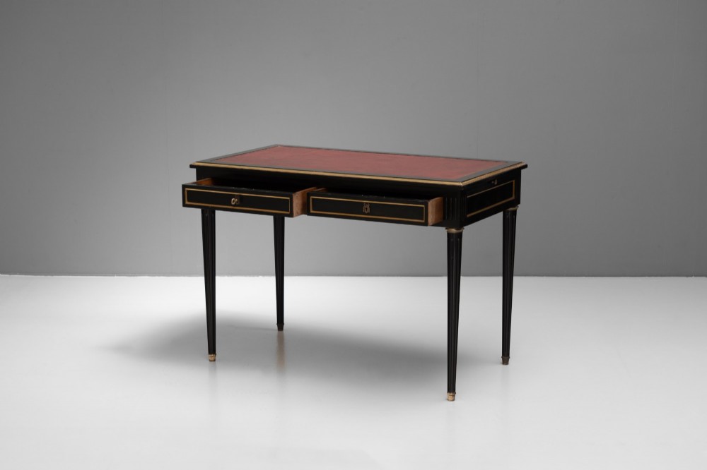 An ebonised wooden writing desk featuring a red leather inset top with gilt brass border inlay, two front drawers, and tapered fluted legs. View V
