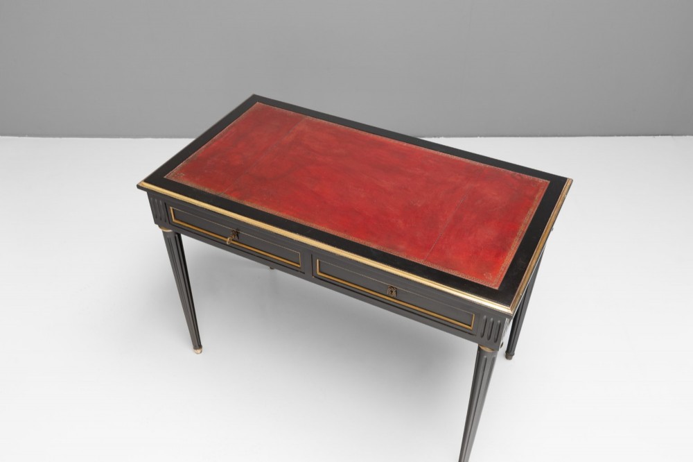 An ebonised wooden writing desk featuring a red leather inset top with gilt brass border inlay, two front drawers, and tapered fluted legs. View VI