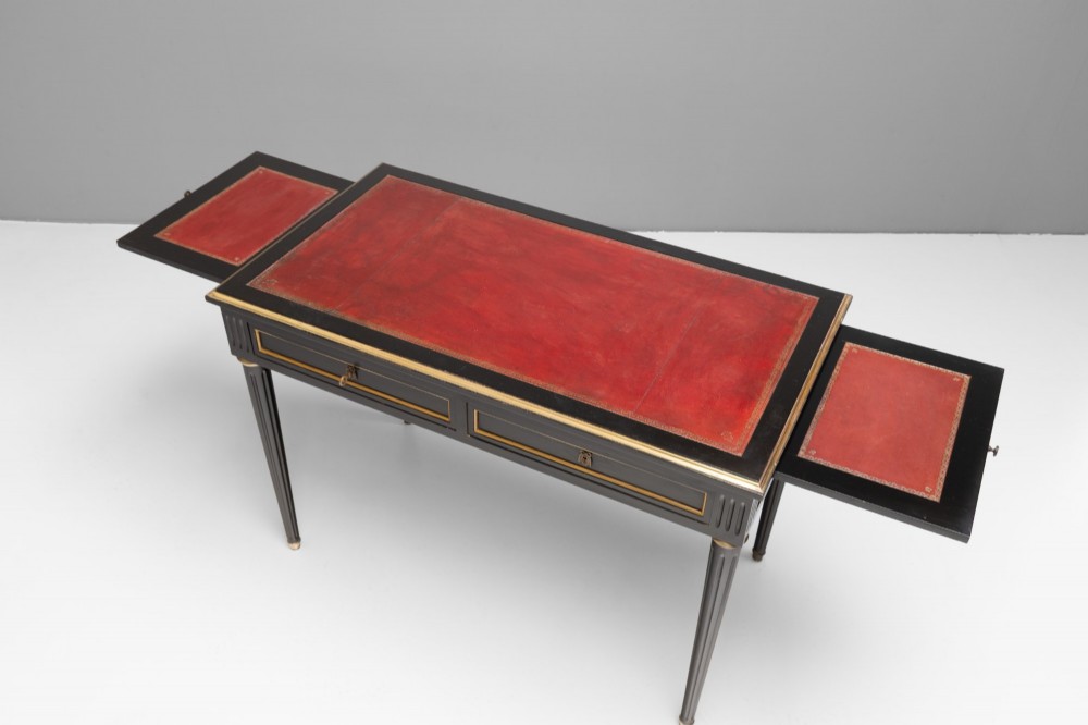An ebonised wooden writing desk featuring a red leather inset top with gilt brass border inlay, two front drawers, and tapered fluted legs. View VII