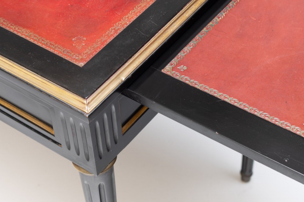 An ebonised wooden writing desk featuring a red leather inset top with gilt brass border inlay, two front drawers, and tapered fluted legs. View VIII