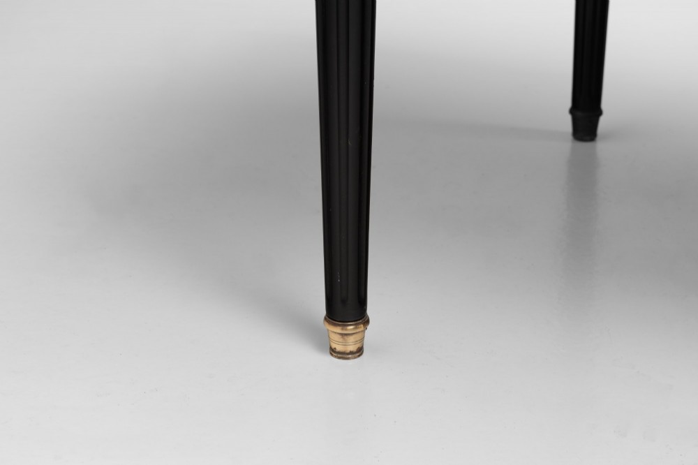 An ebonised wooden writing desk featuring a red leather inset top with gilt brass border inlay, two front drawers, and tapered fluted legs. View X