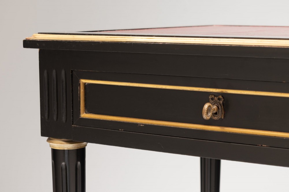 An ebonised wooden writing desk featuring a red leather inset top with gilt brass border inlay, two front drawers, and tapered fluted legs. View XI