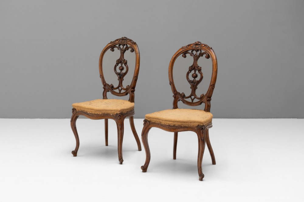 A pair of antique Victorian Rococo Revival carved timber side chairs with balloon backs, open scrollwork splats, cabriole legs, and neutral upholstery.