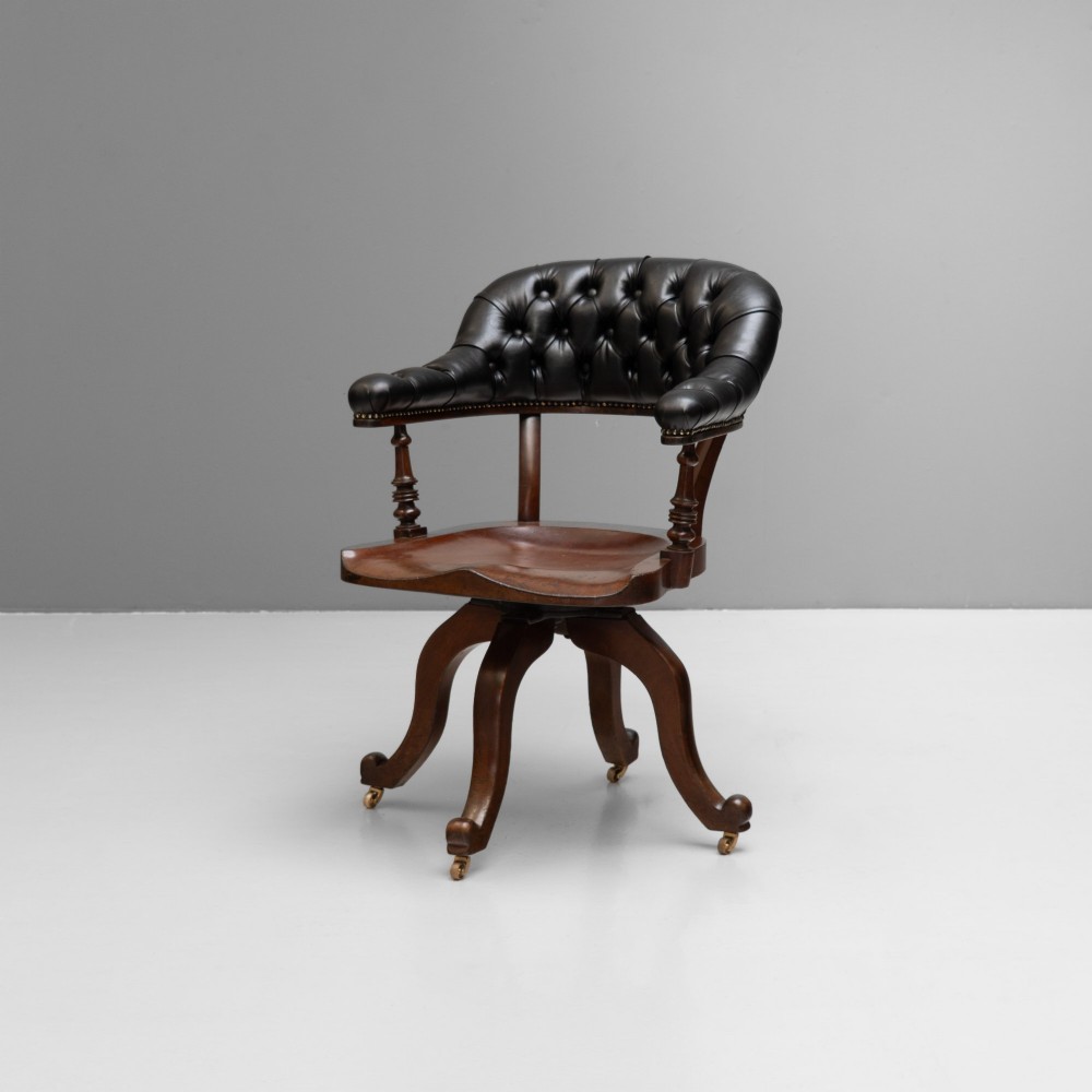 An antique Victorian swivel desk chair featuring a deep-buttoned dark leather backrest, shaped wooden seat, turned arm supports, and splayed legs on brass castors.