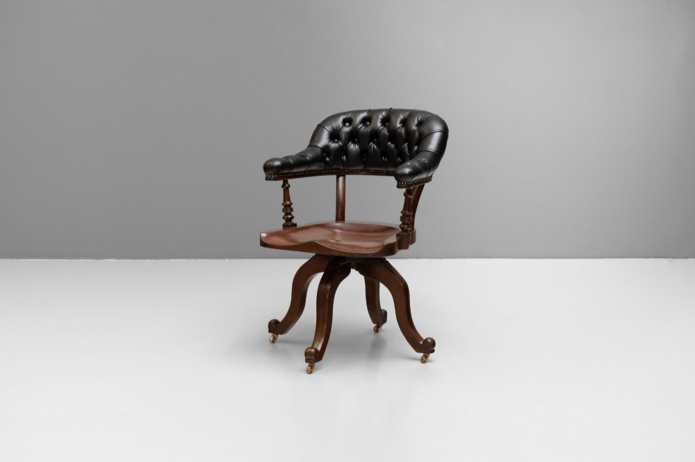 An antique Victorian swivel desk chair featuring a deep-buttoned dark leather backrest, shaped wooden seat, turned arm supports, and splayed legs on brass castors. View II