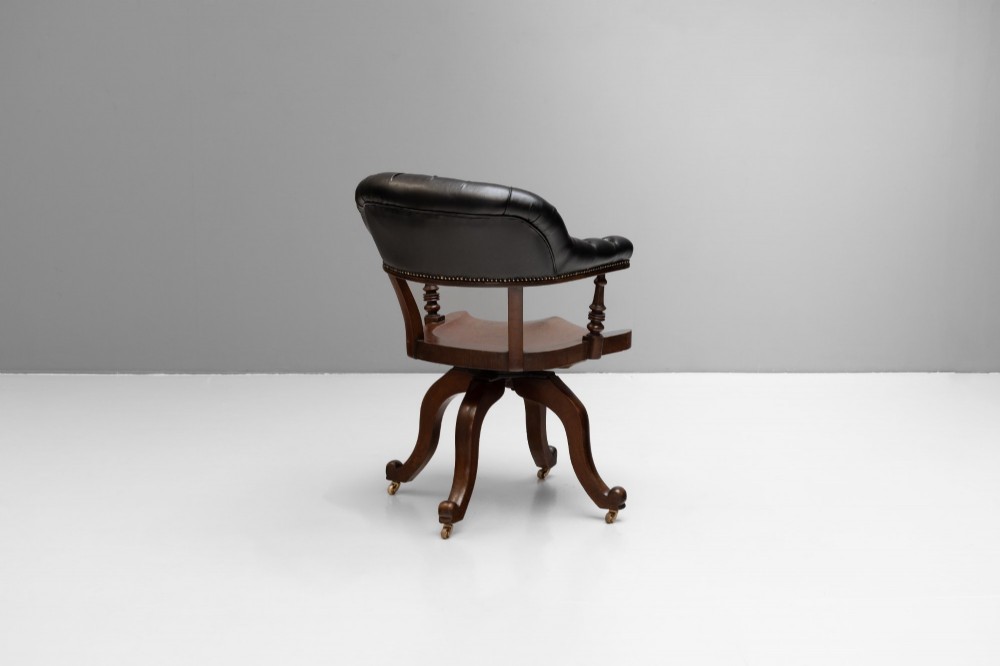 An antique Victorian swivel desk chair featuring a deep-buttoned dark leather backrest, shaped wooden seat, turned arm supports, and splayed legs on brass castors. View III