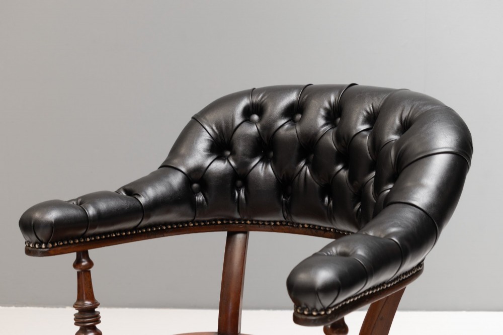 An antique Victorian swivel desk chair featuring a deep-buttoned dark leather backrest, shaped wooden seat, turned arm supports, and splayed legs on brass castors. View IV
