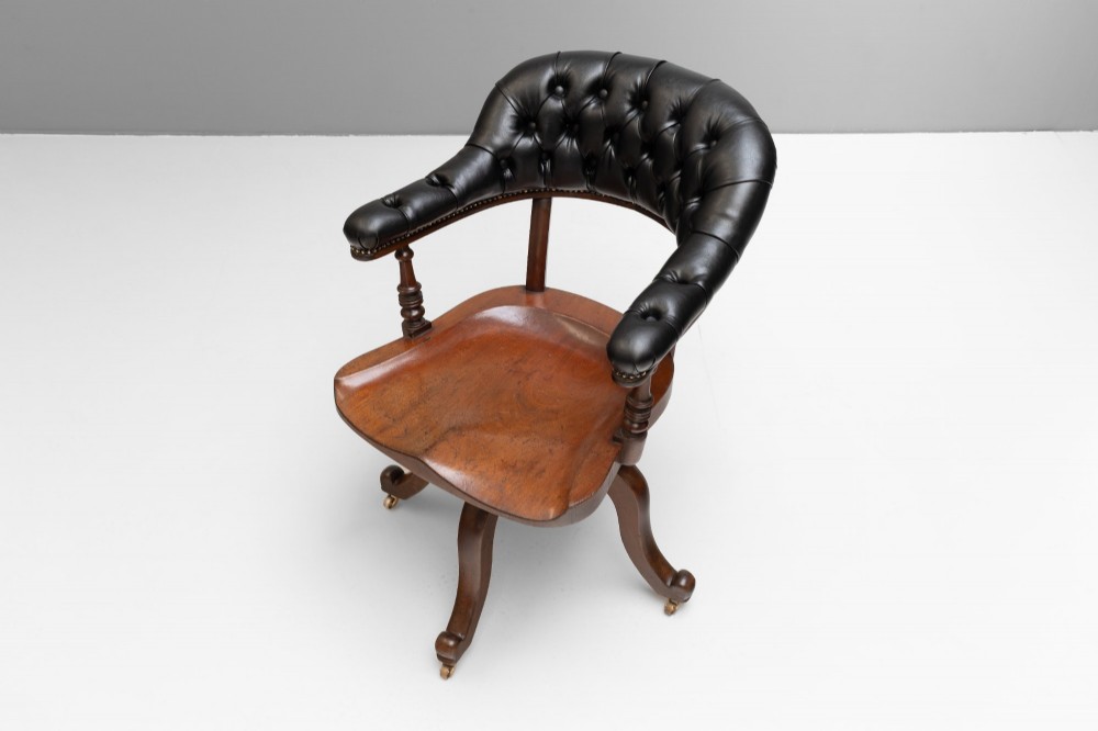 An antique Victorian swivel desk chair featuring a deep-buttoned dark leather backrest, shaped wooden seat, turned arm supports, and splayed legs on brass castors. View VI