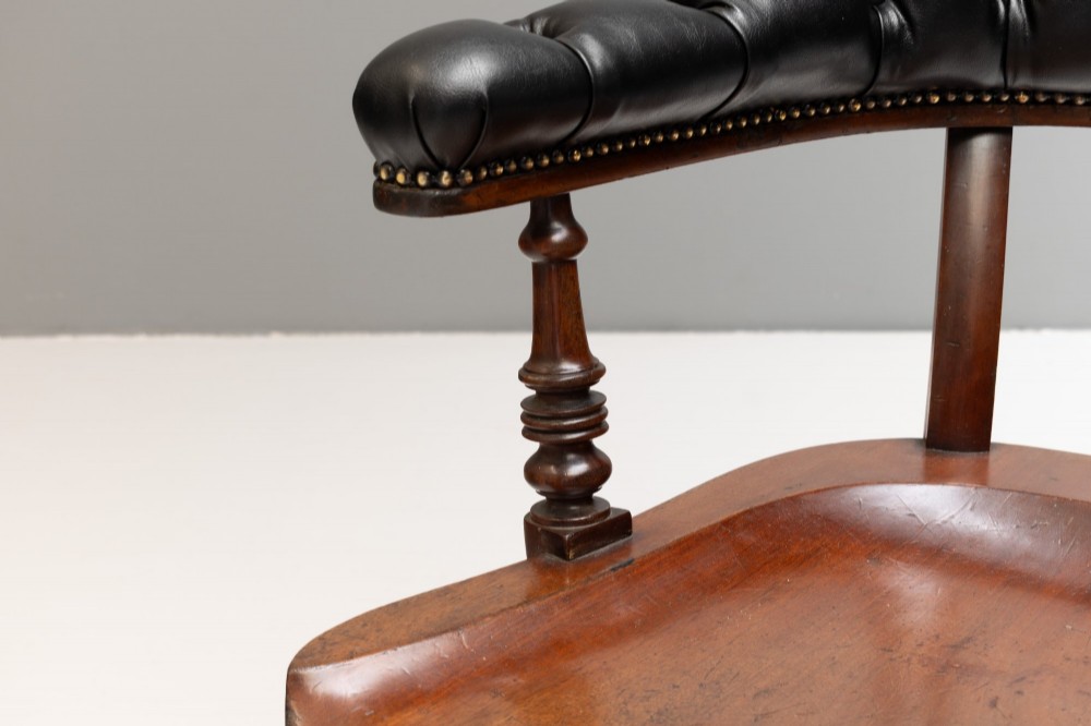 An antique Victorian swivel desk chair featuring a deep-buttoned dark leather backrest, shaped wooden seat, turned arm supports, and splayed legs on brass castors. View VII