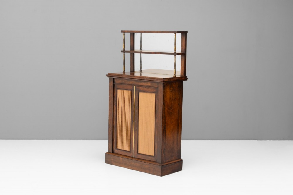 An antique rosewood chiffonier featuring a two-tier mirrored shelf superstructure supported by brass columns over a double-door cupboard with fabric panels.