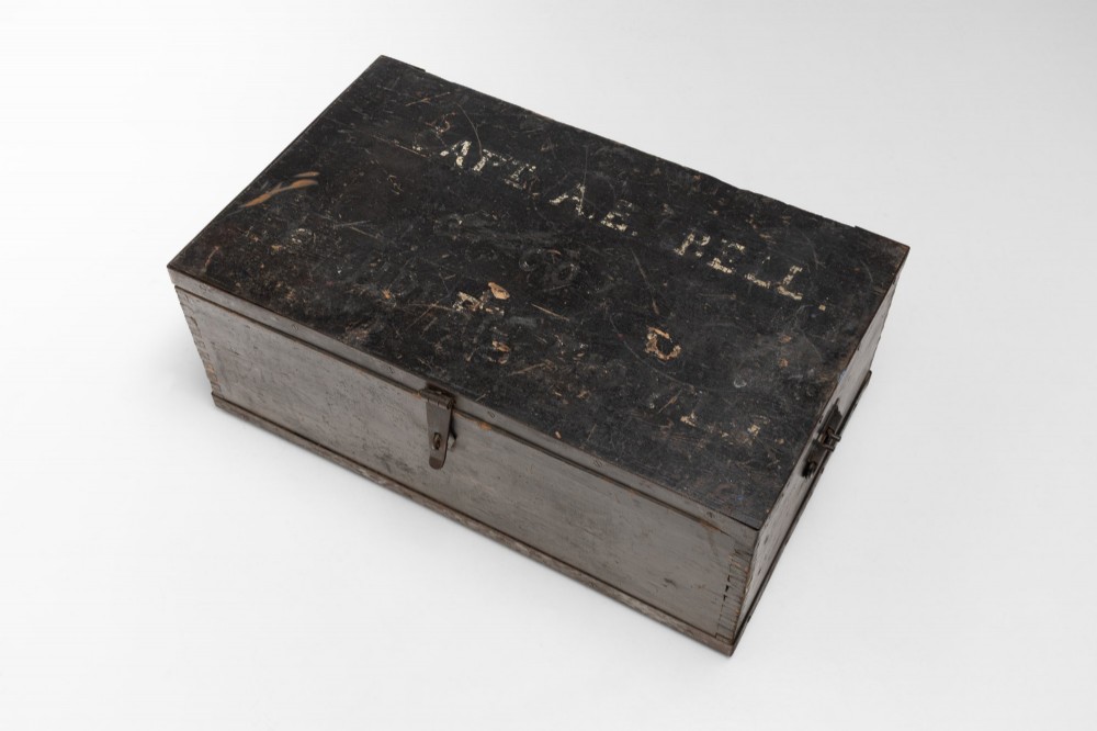 A dark brown rectangular wooden sea chest featuring white painted lettering on the flat lid and iron drop side handles. View III