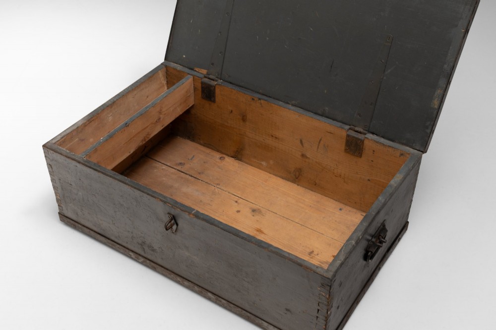 A dark brown rectangular wooden sea chest featuring white painted lettering on the flat lid and iron drop side handles. View V