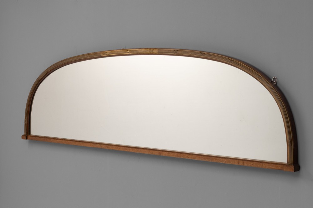 A wide arched overmantel mirror featuring a reeded gilt frame with detailed cross-ribbon motifs and a flat wooden base.