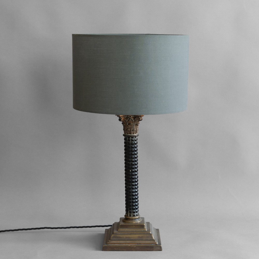 A Neoclassical style table lamp with a dark textured column shaft, an ornate brass Corinthian capital, a stepped brass base, and a grey cylindrical drum shade.
