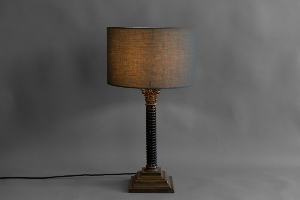 A Neoclassical style table lamp with a dark textured column shaft, an ornate brass Corinthian capital, a stepped brass base, and a grey cylindrical drum shade. View II