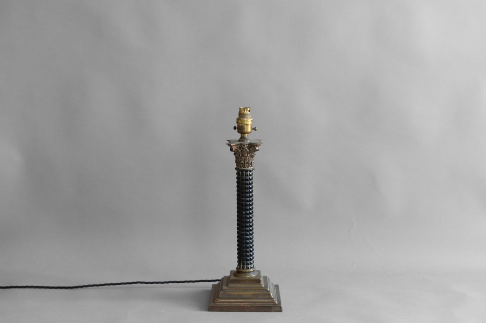 A Neoclassical style table lamp with a dark textured column shaft, an ornate brass Corinthian capital, a stepped brass base, and a grey cylindrical drum shade. View III