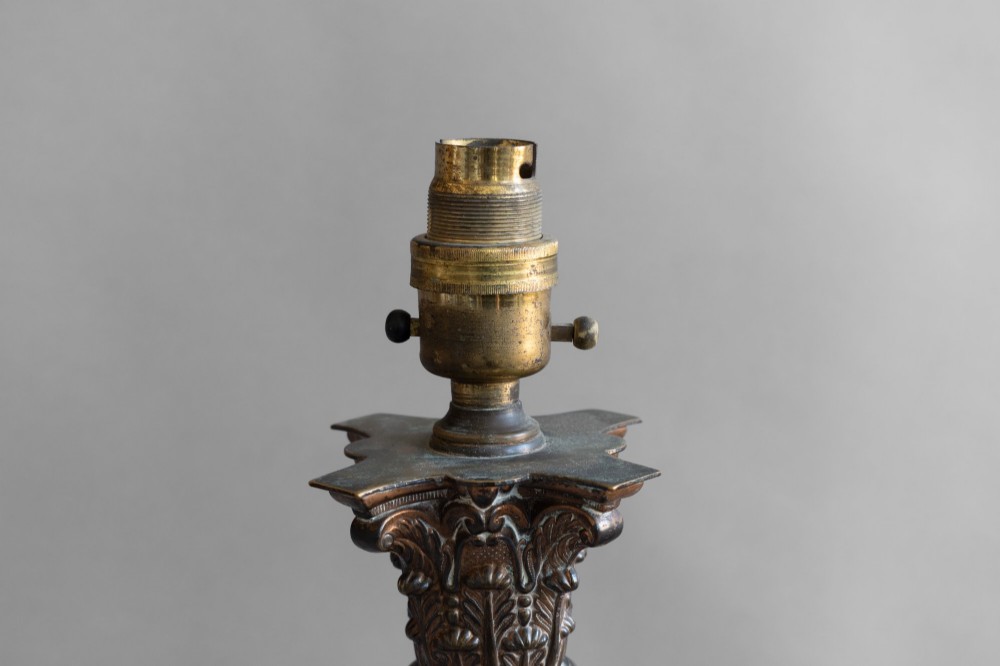 A Neoclassical style table lamp with a dark textured column shaft, an ornate brass Corinthian capital, a stepped brass base, and a grey cylindrical drum shade. View IV
