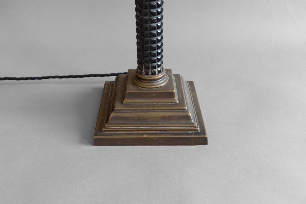 A Neoclassical style table lamp with a dark textured column shaft, an ornate brass Corinthian capital, a stepped brass base, and a grey cylindrical drum shade. View VI