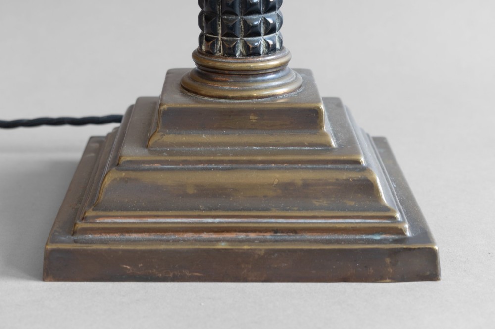 A Neoclassical style table lamp with a dark textured column shaft, an ornate brass Corinthian capital, a stepped brass base, and a grey cylindrical drum shade. View VII