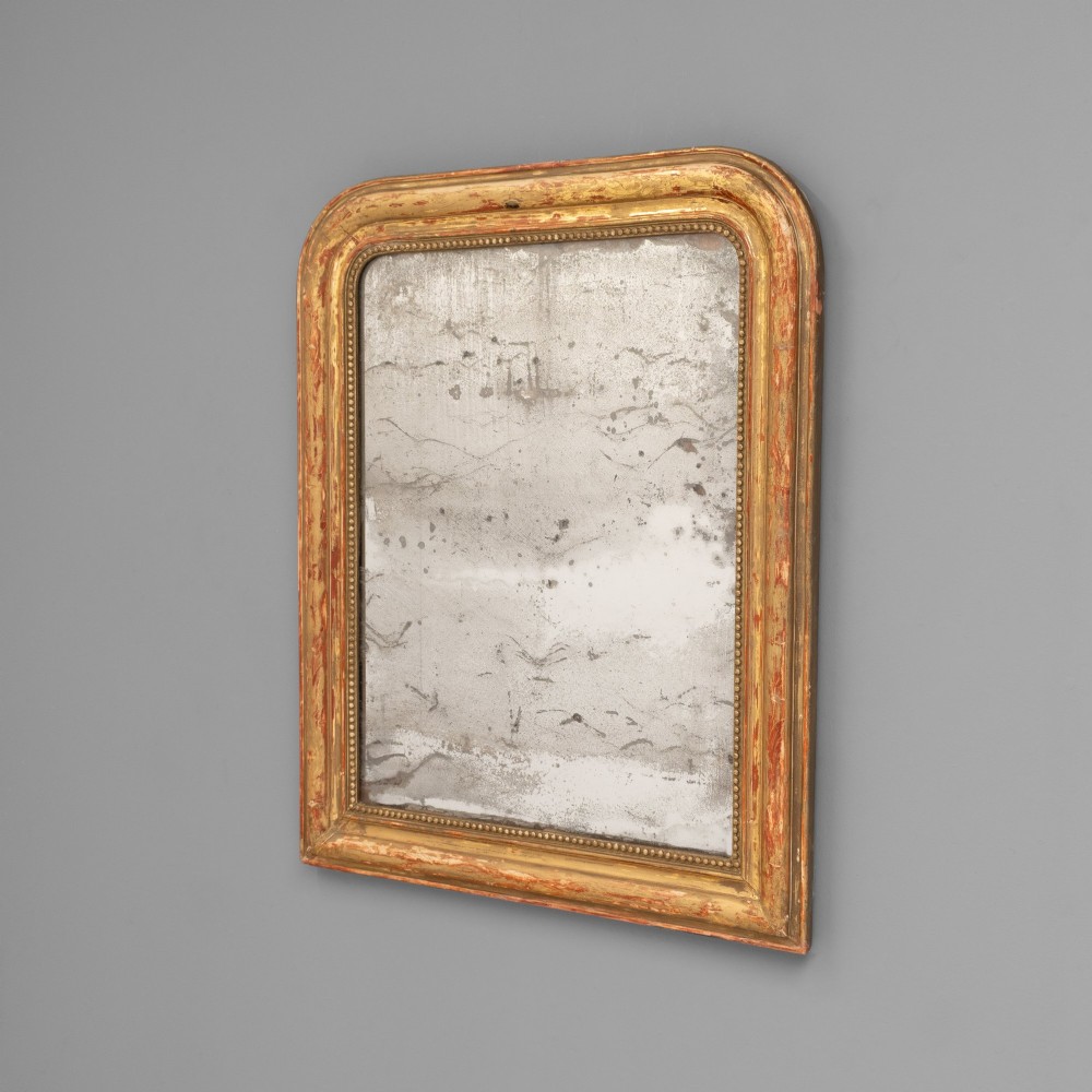 A Louis Philippe style wall mirror featuring a rounded rectangular giltwood frame with visible red bole ground, beaded inner trim, and heavily foxed glass.