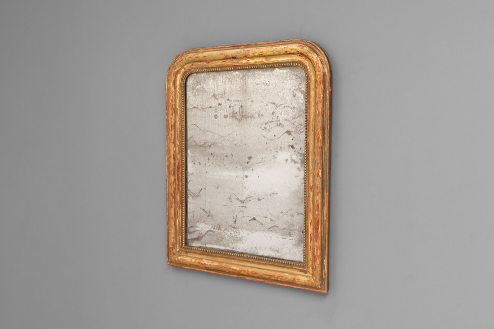 A Louis Philippe style wall mirror featuring a rounded rectangular giltwood frame with visible red bole ground, beaded inner trim, and heavily foxed glass. View II