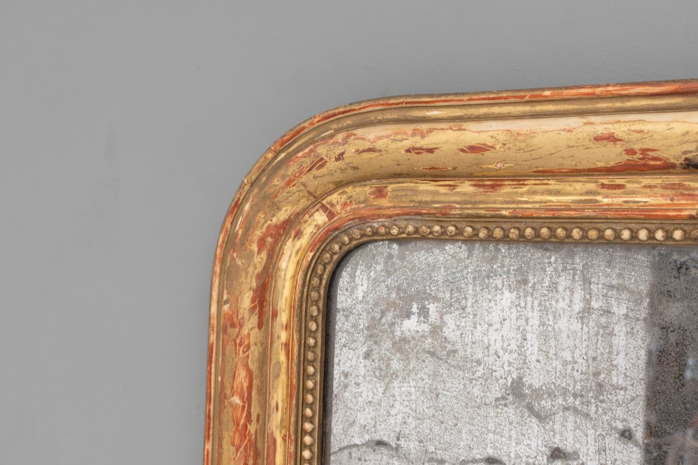 A Louis Philippe style wall mirror featuring a rounded rectangular giltwood frame with visible red bole ground, beaded inner trim, and heavily foxed glass. View III