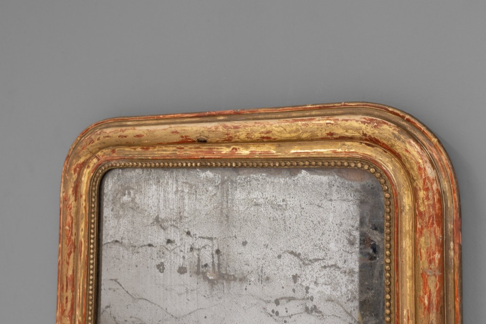 A Louis Philippe style wall mirror featuring a rounded rectangular giltwood frame with visible red bole ground, beaded inner trim, and heavily foxed glass. View IV